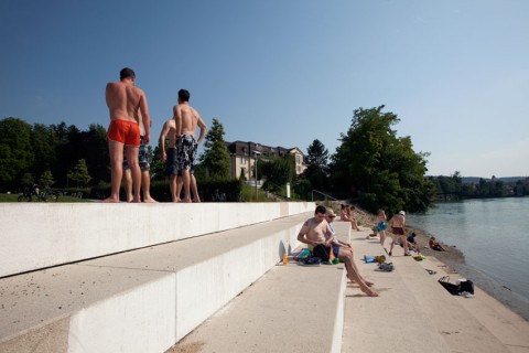 Menschen sitzen oder stehen auf in Badehosen auf breiten Stufen, welche zum Fluss führen