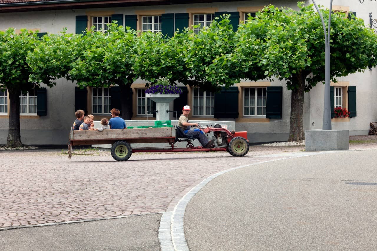 Kinder fahren auf der Ladefläche eines landwirtschaftlichen Fahrzeugs über den Dorfplatz von Wölflinswil.