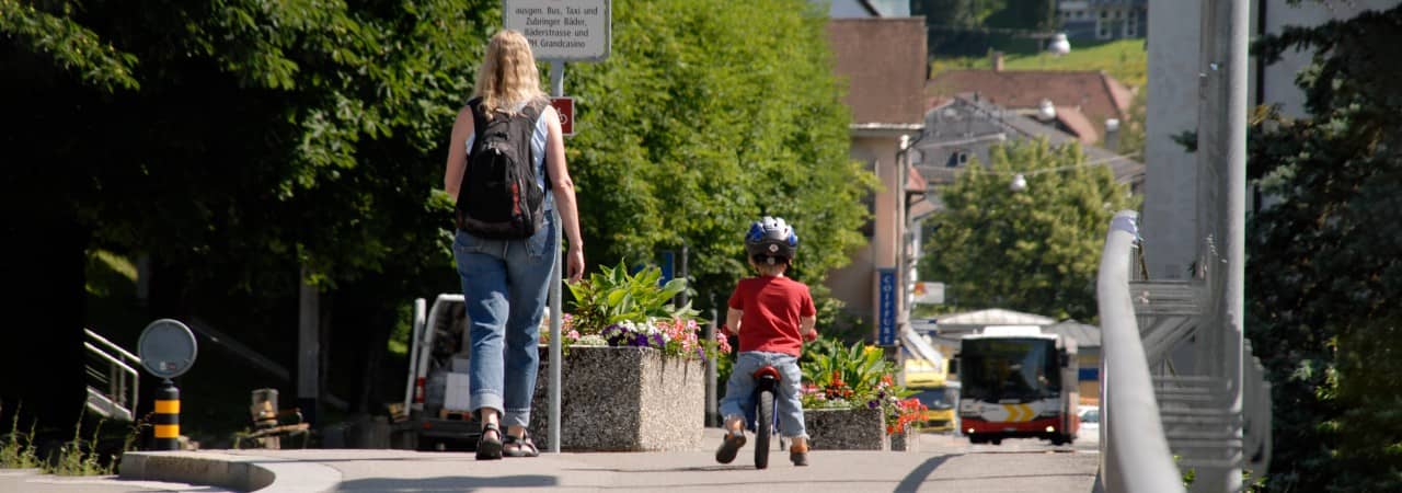 Foto einer Fussgängerin, eines Kindes mit Velo und einem Bus im Hintergrund auf einer Strasse in Baden.