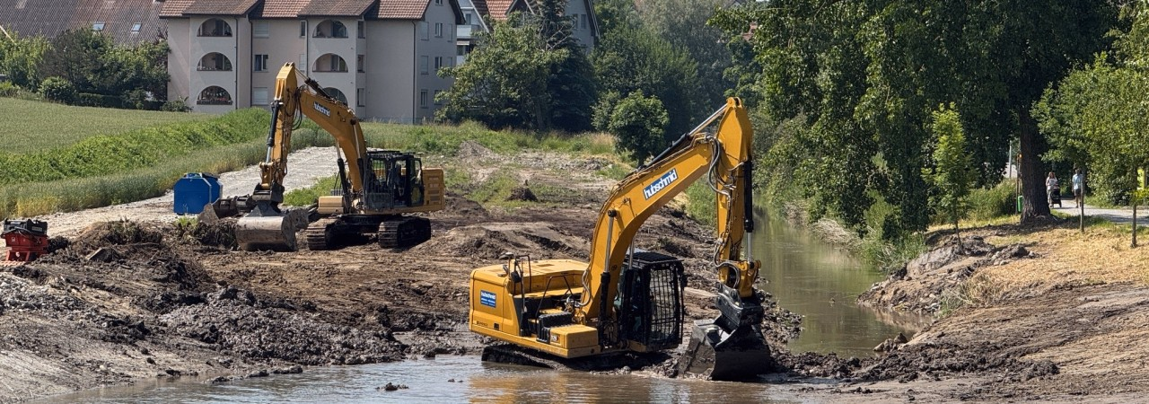 Zwei Bagger führen Arbeiten in einem flachen Gewässer durch, umgeben von trockener Erdmasse. Im Hintergrund sind Wohnhäuser, Bäume und ein Feld erkennbar.
