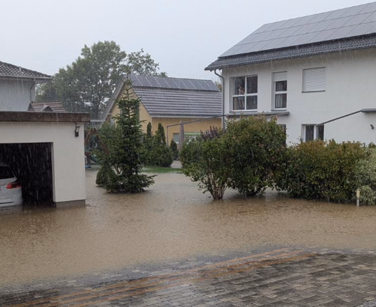 Überschwemmte Wohnsiedlung mit mehreren Häusern, Garagen und Solarpaneelen auf den Dächern, umgeben von teils bepflanzten Vorgärten.