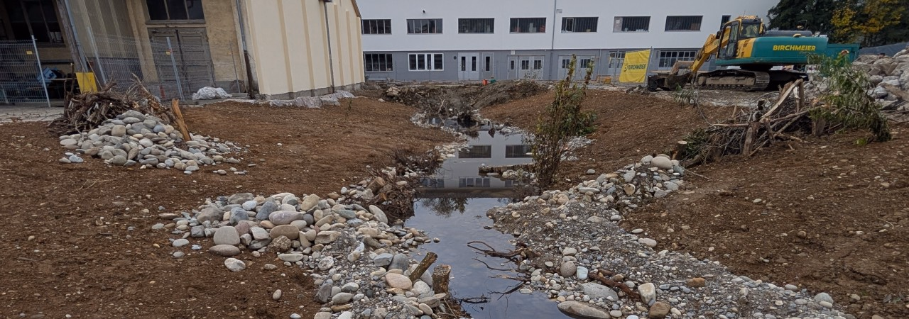 Unbebautes Gelände mit einem kleinen Bachlauf aus Kiessteinen, einem gelben Bagger rechts und Gebäuden im Hintergrund.