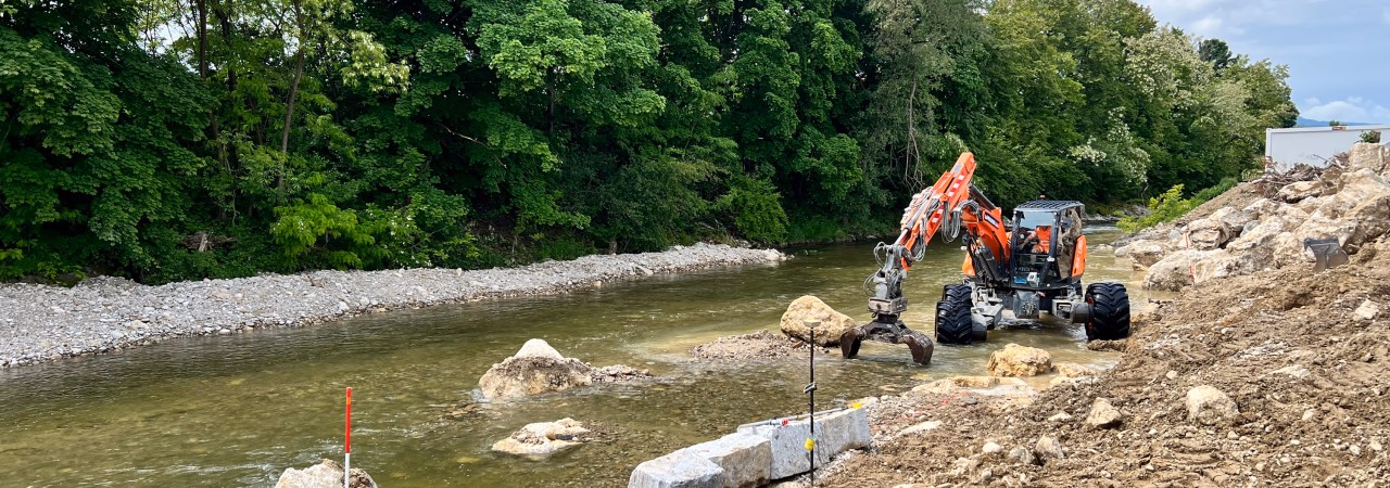 Ein Bagger arbeitet am Ufer eines Flusses, umgeben von Kies, Steinen und Messtechnik. Im Hintergrund sind Bäume und eine helle Wolkendecke.
