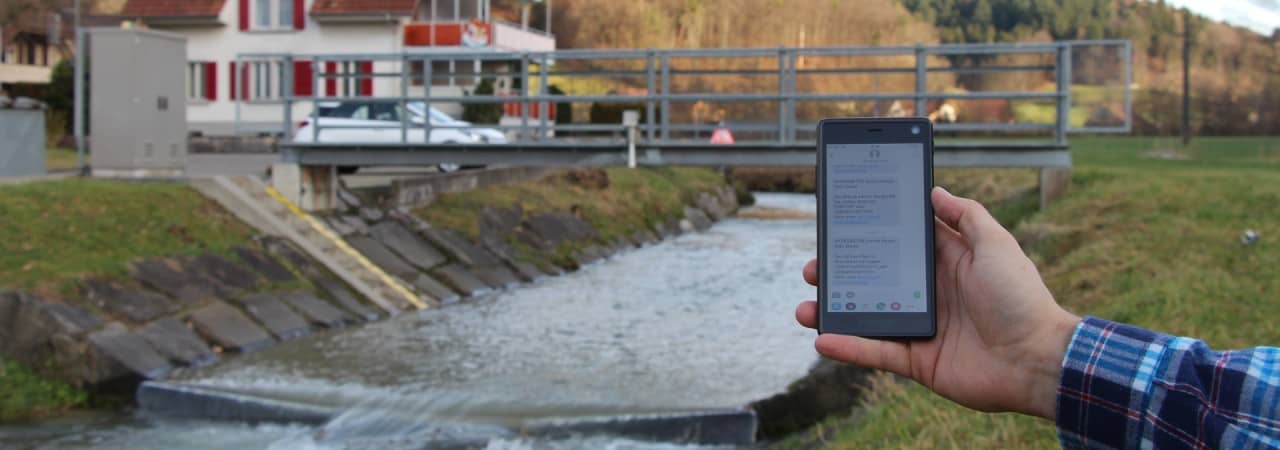 Eine Hand hält ein Smartphone auf dessen Bildschirm SMS zu sehen sind. Im Hintergrund ist eine Brücke über einen Bach zu sehen; an der Brücke ist eine Messstation installiert.