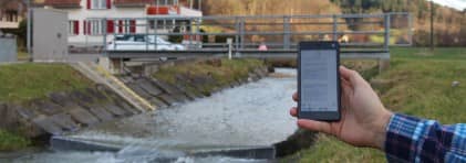 Eine Hand hält ein Smartphone auf dessen Bildschirm SMS zu sehen sind. Im Hintergrund ist eine Brücke über einen Bach zu sehen; an der Brücke ist eine Messstation installiert.