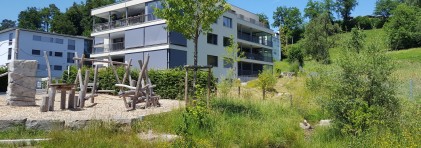 Das Bild zeigt ein Wohnhaus am Siedlungsrand mit einem kleinen Wiesenbach im Vordergrund