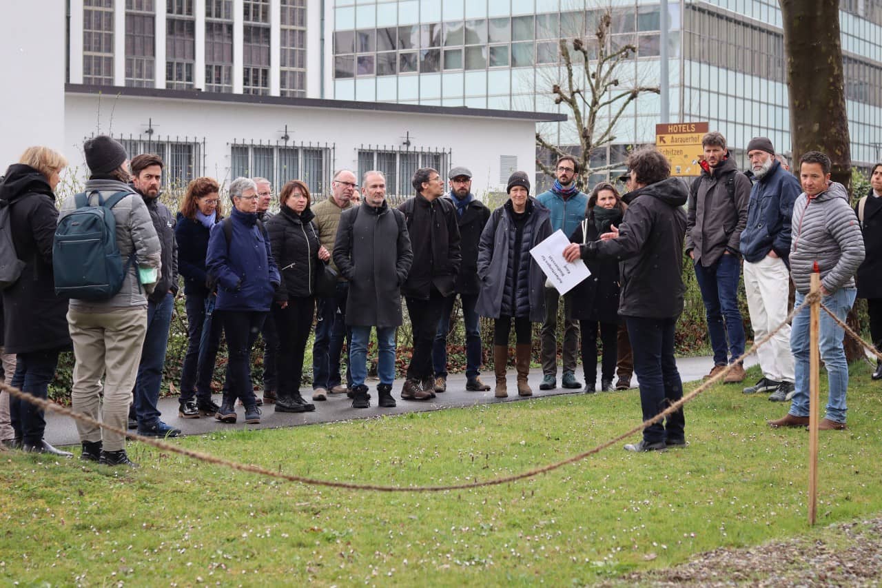 Ein Publikum hört einer Person zu, die draussen einen Vortrag hält.