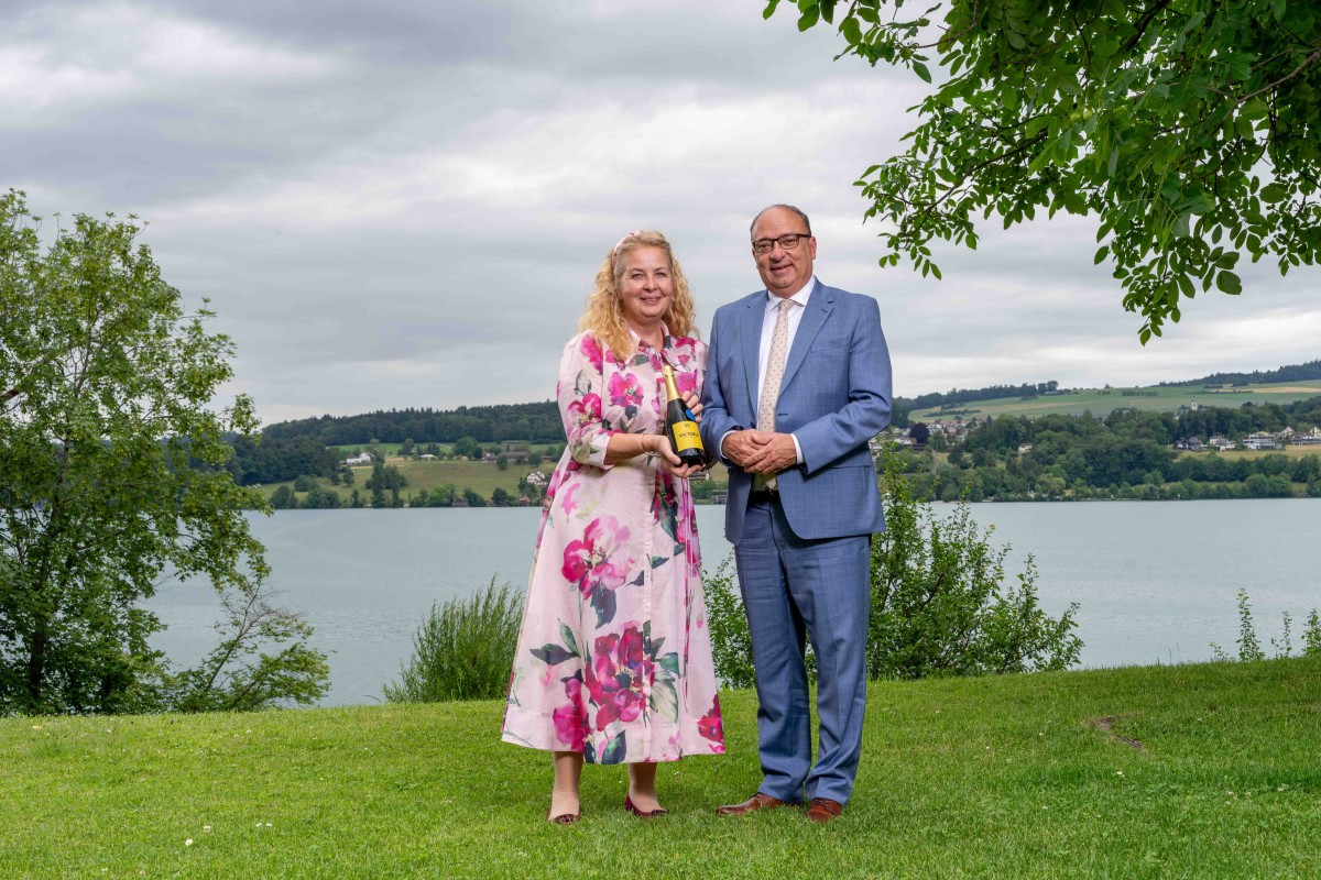 Zwei Personen stehen vor einem See und präsentieren eine Flasche Wein.