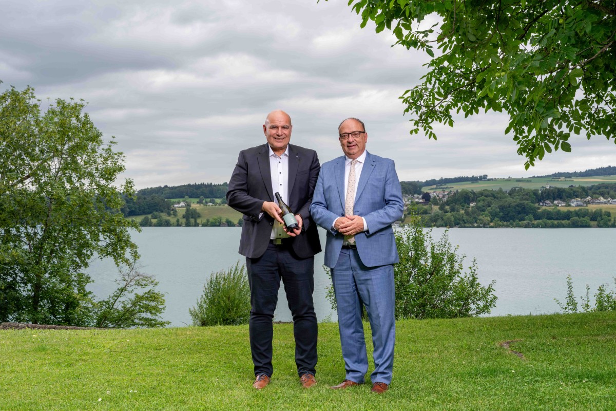 Zwei Personen stehen vor einem See und präsentieren eine Flasche Wein.
