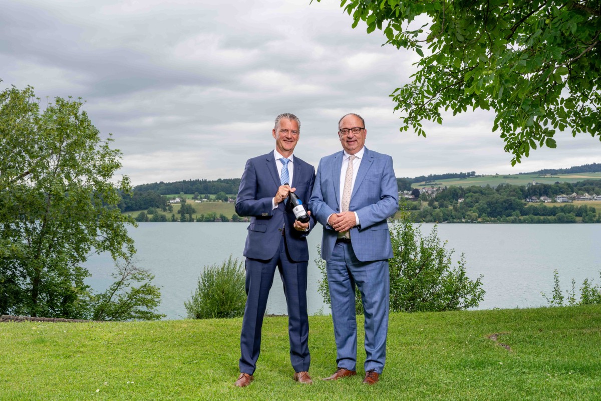 Zwei Personen stehen vor einem See und präsentieren eine Flasche Wein.