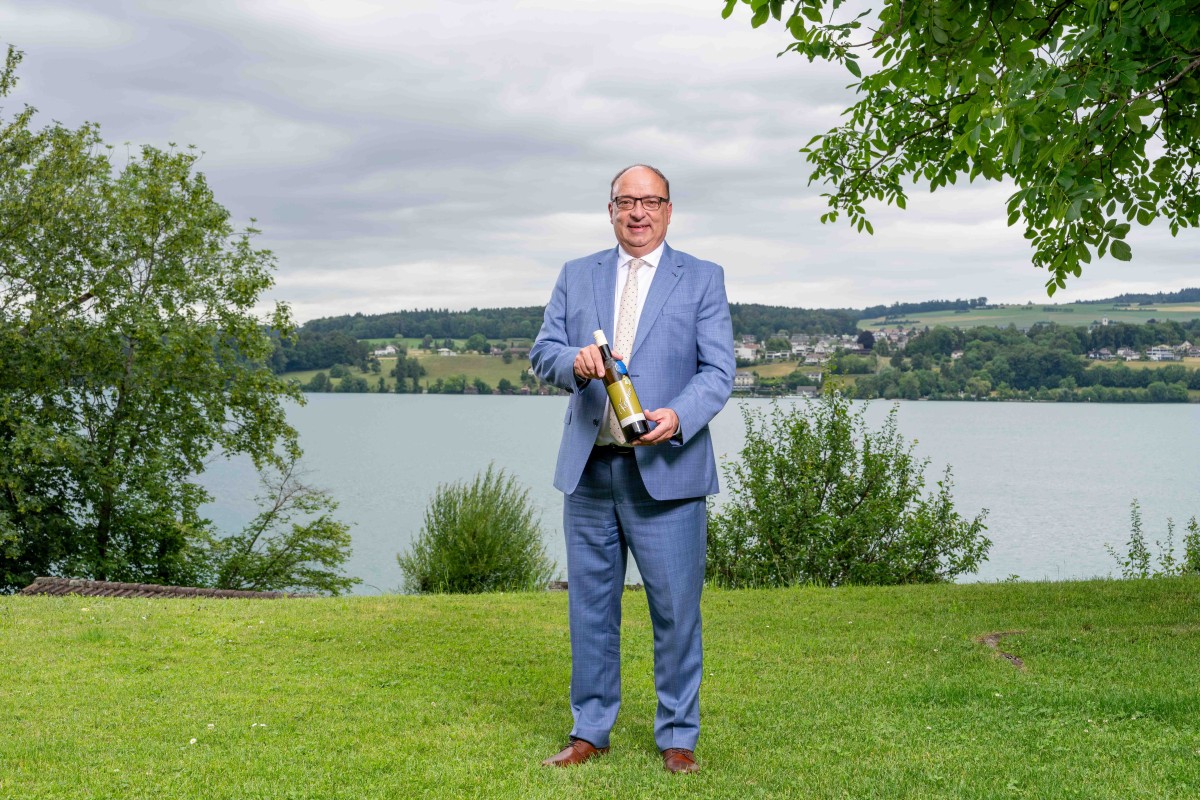 Ein Mann steht vor einem See und präsentiert eine Flasche Wein.