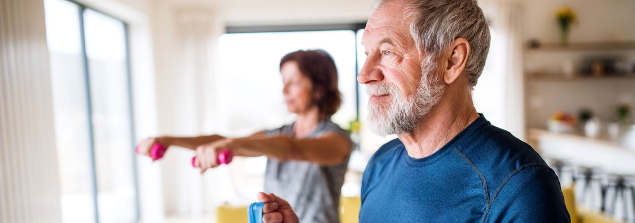Senior mit Theraband im Vordergrund, Seniorin im Hintergrund mit zwei Hanteln.