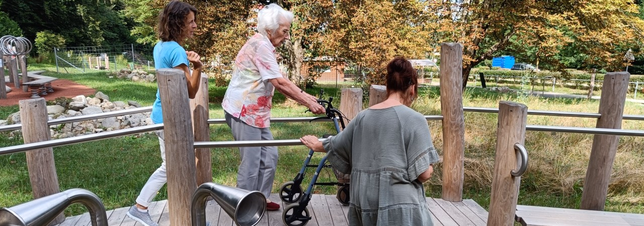 Ältere Frau an Rollator läuft über einen Holzweg, zwei jüngere Frauen stehen hinter/neben ihr.