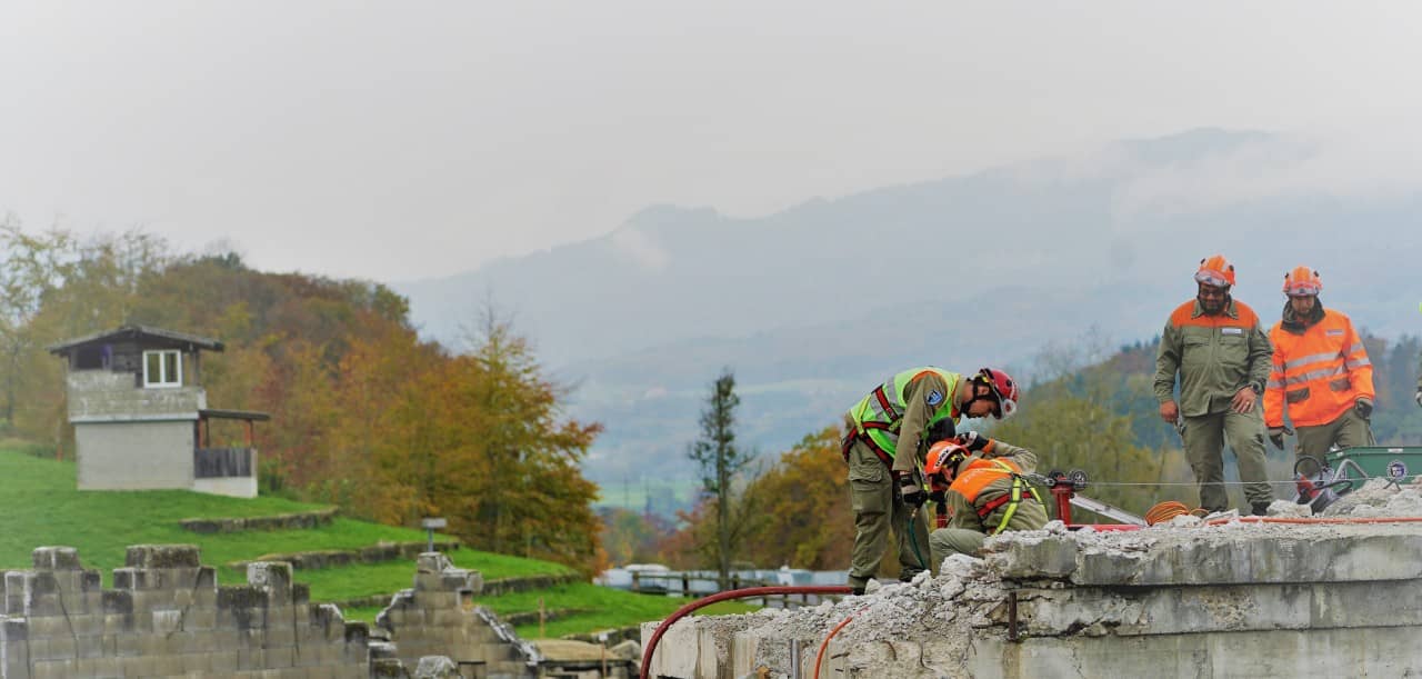 Zivilschützer bereiten eine Kernbohrung vor. Im Hintergrund sieht man ein Haus des Übungsdorfes.