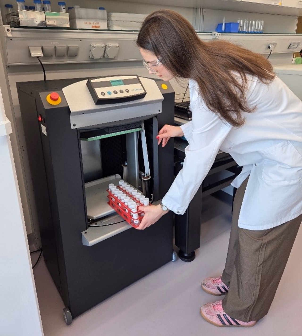 Person in weissem Laborkittel mit Laborbrille stellt ein Gefäss mit Röhrchen in eine Labormaschine.