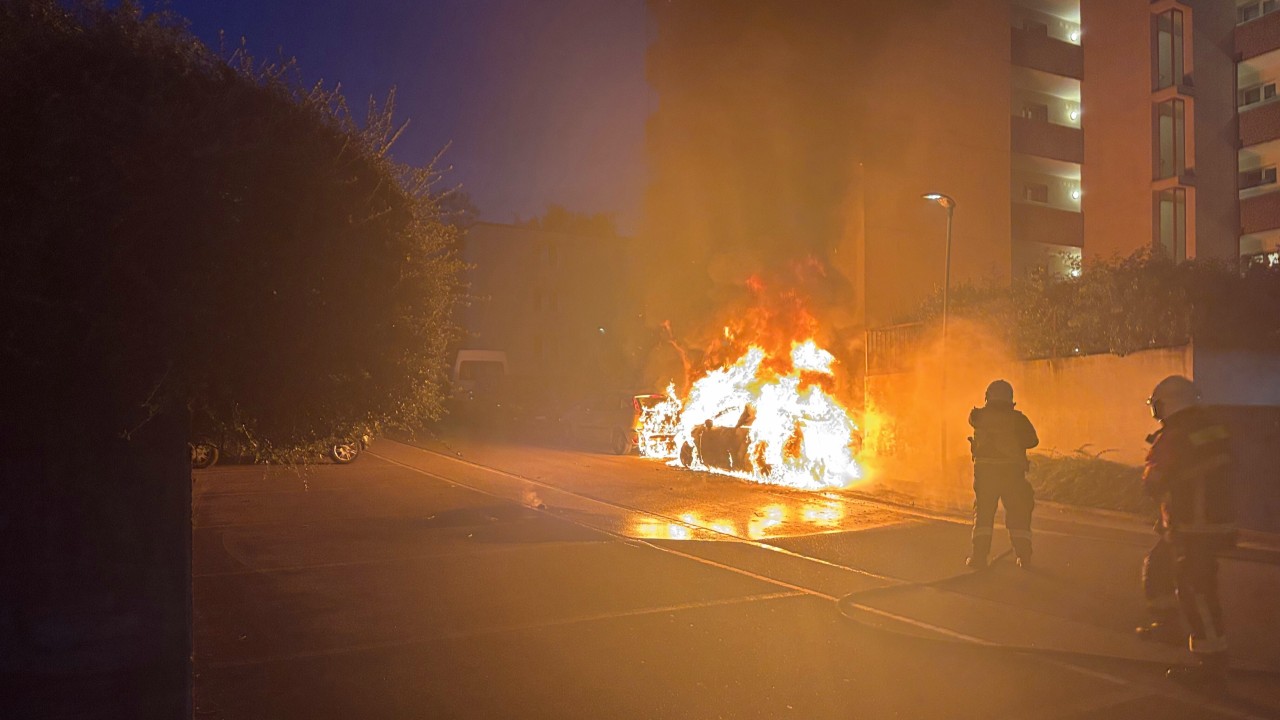 Brennendes Auto bei Nacht auf einer Strasse, waehrend zwei Feuerwehrleute den Brand loeschen.
