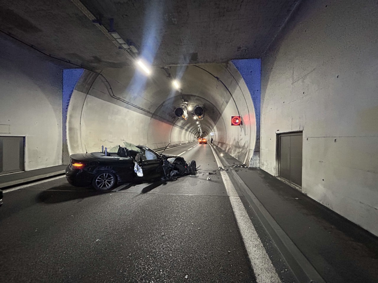 Schwarzes BMW Cabriolet, in einem Tunnel. Die Front ist stark beschädigt. Weiter vorne ein weiteres Fahrzeug stillstehend im Tunnel.