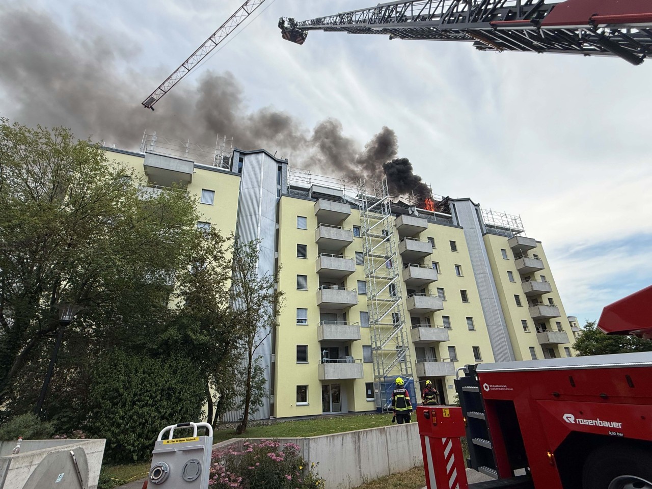 Auf dem Dach eines Mehrfamilienhauses ist ein Brand ersichtlich. Feuerwehr mit Drehleiter vor Ort.