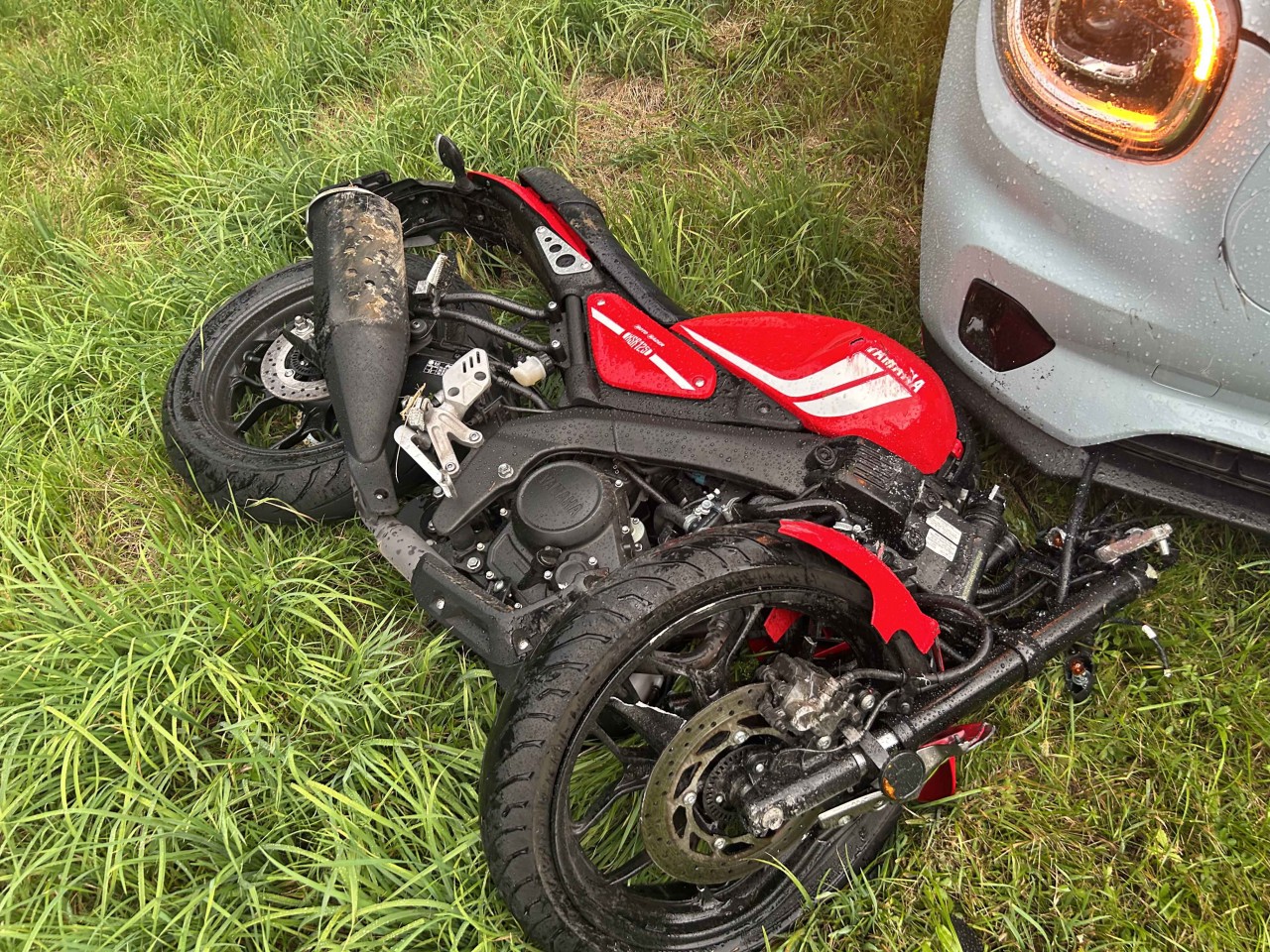Beschädigtes, rotes Motorrad im Gras liegend vor einer Autofront.