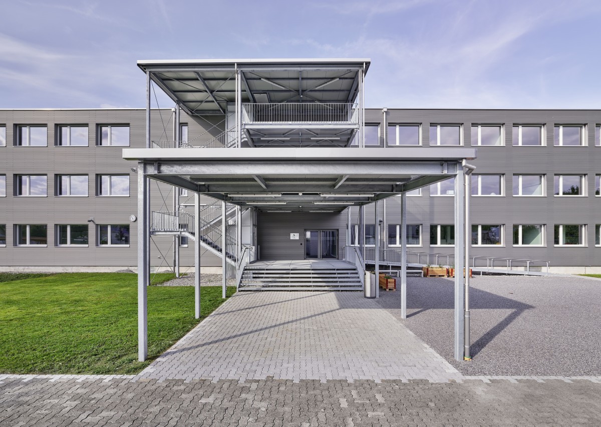 Das Bild zeigt den überdachten Eingang des Modulbaus der Kantonsschule Stein. Das Gebäude ist aus grauem Holz. Links vom Eingang sieht man eine grüne Wiese, rechts davcon einen Platz mit Kieselsteinen.