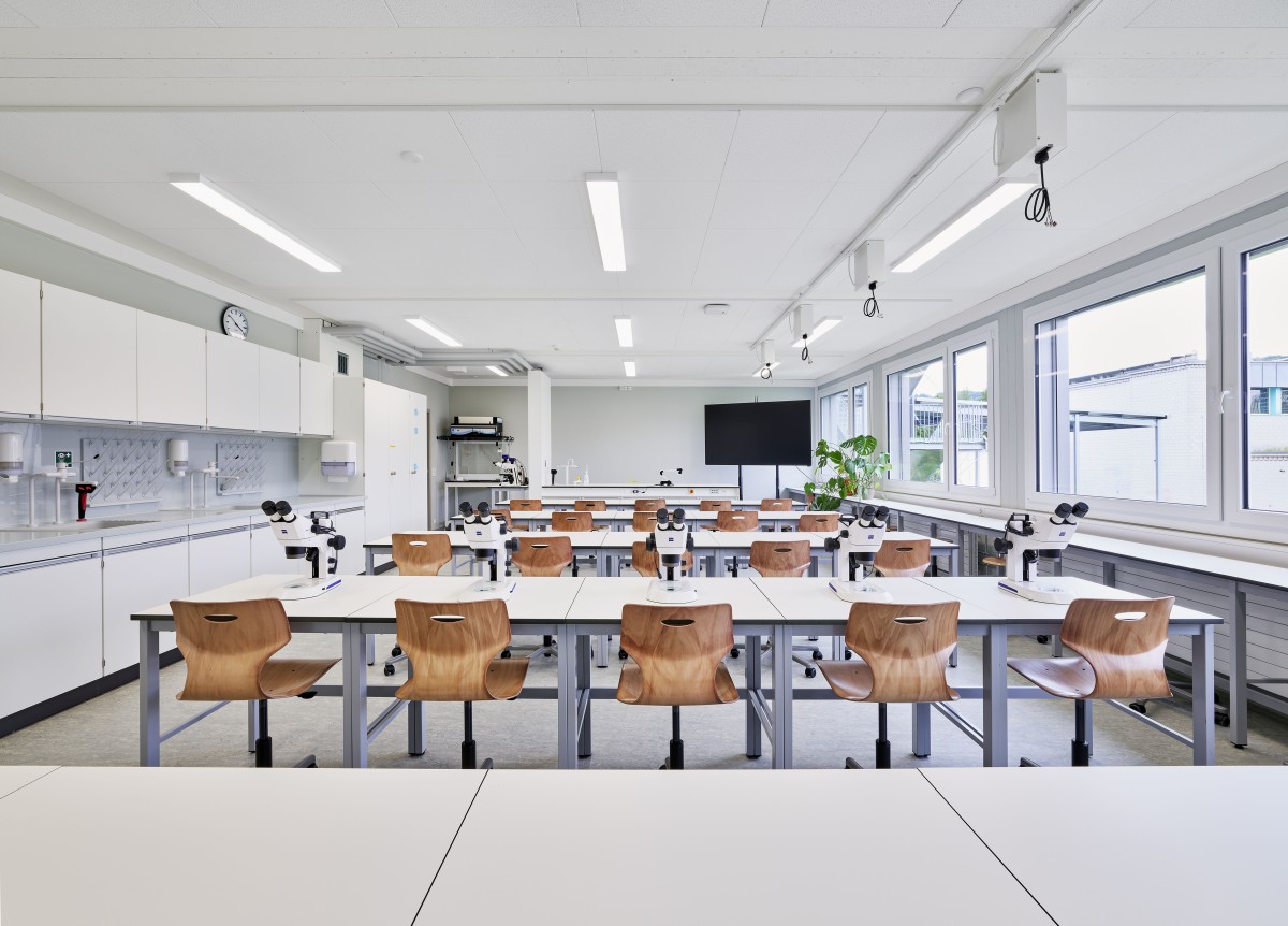 Auf dem Bild ist ein modernes Schulzimmer zu sehen, mit weissen Tischen und holzigen Stühlen. Auf einer Tischreihe stehen Mikroskope, eine grüne Pflanze steht hinten rechts in der Ecke.