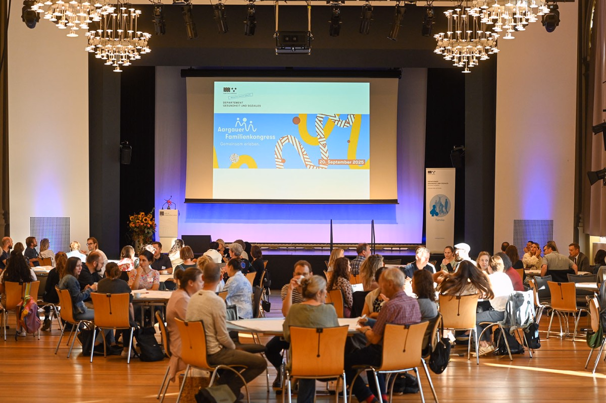 Halle mit mehreren Tischen mit Menschen, die zu einer Leinwand mit Präsentetion schauen.