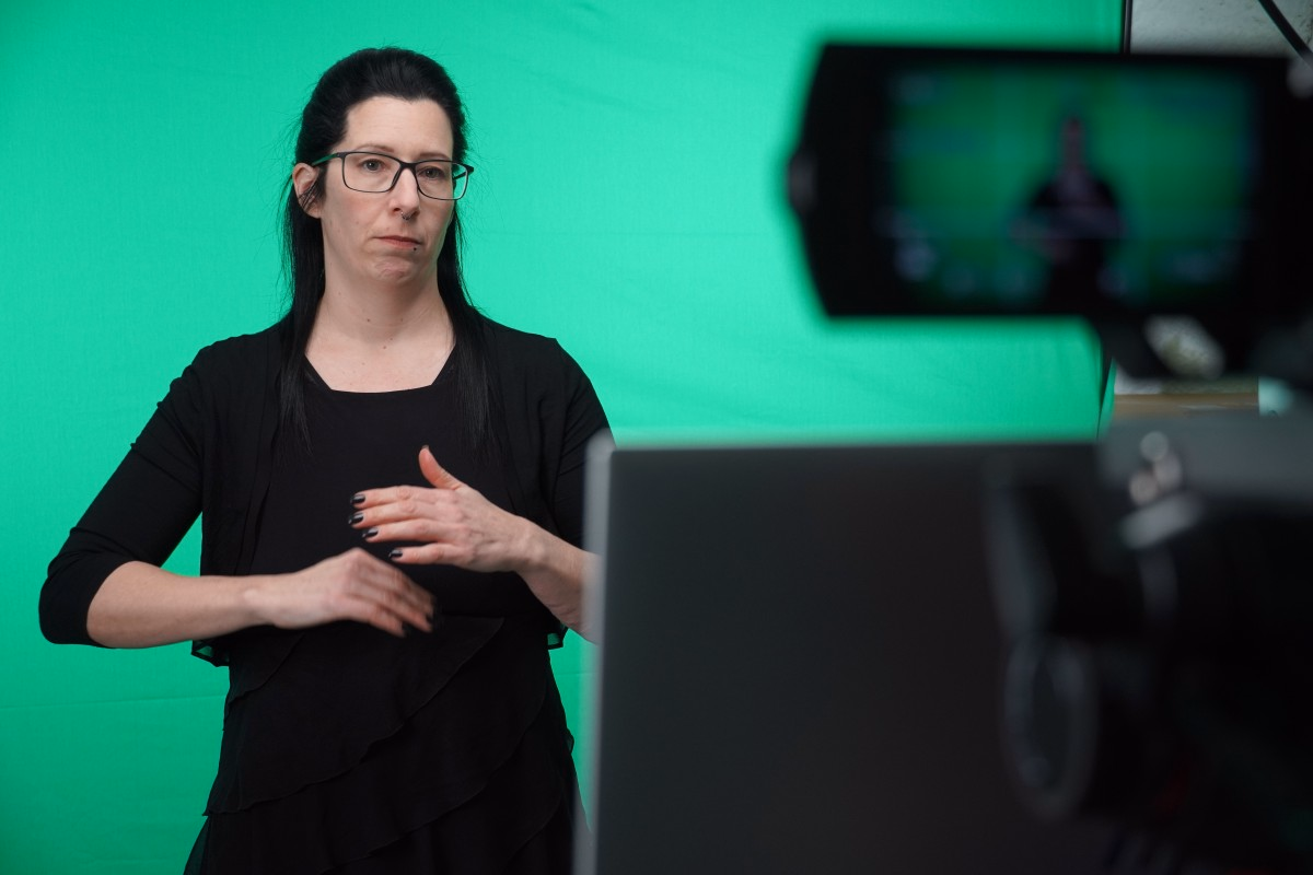 Eine Frau mit dunklen Haaren und schwarzer Kleidung steht vor einem grünen Hintergrund und spricht vor einer Kamera.