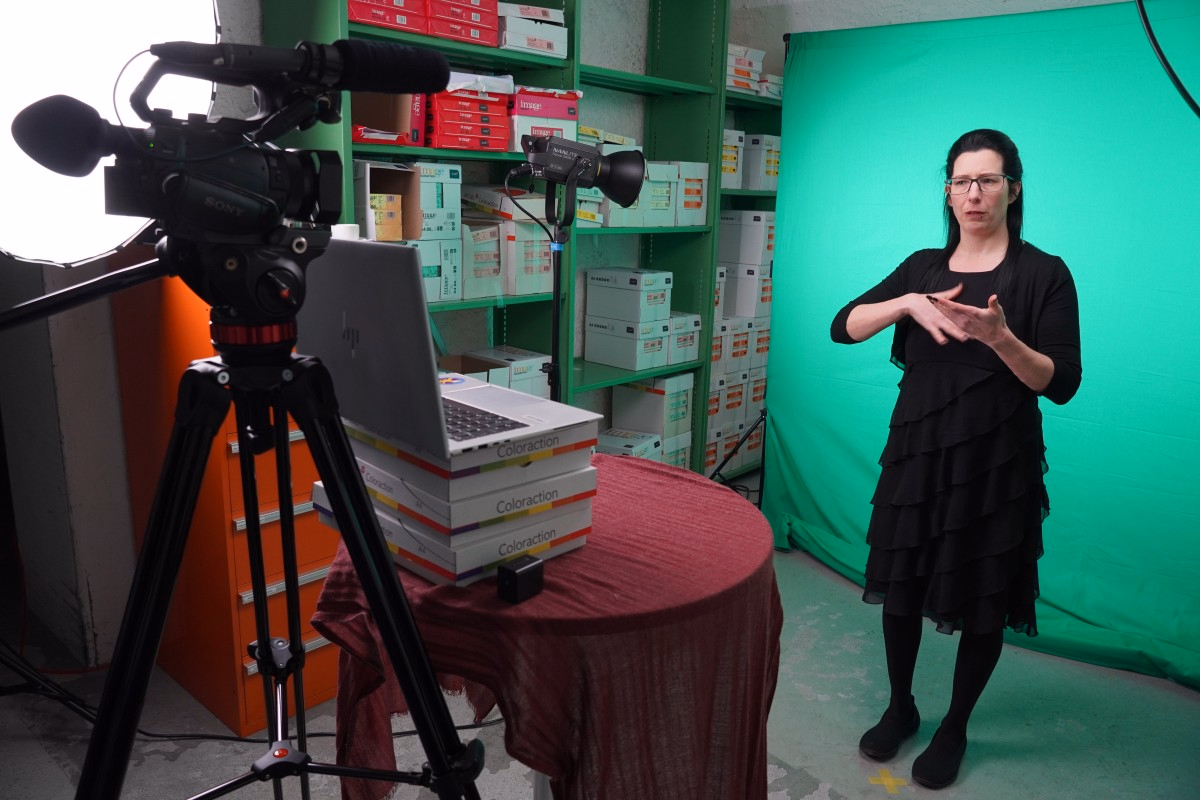 Eine Frau mit dunklen Haaren und schwarzer Kleidung steht vor einer grünen Wand, einer Kamera und einem Tisch mit Büchern und Laptop. Im Hintergrund ist ein Gestell mit Büromaterialien zu sehen.