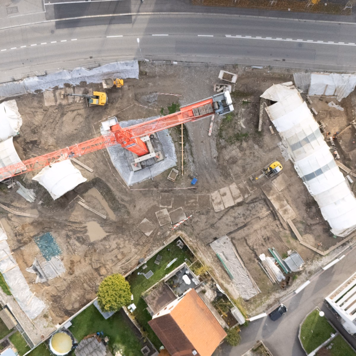 Drohnenaufnahme einer grossen Baustelle inmitten eines Wohnquartiers: Ein roter Turmdrehkran überspannt das Gelände, mehrere weiss abgedeckte Gräben und Erdhügel sind zu erkennen, oben verläuft eine Strasse mit einzelnen Autos.