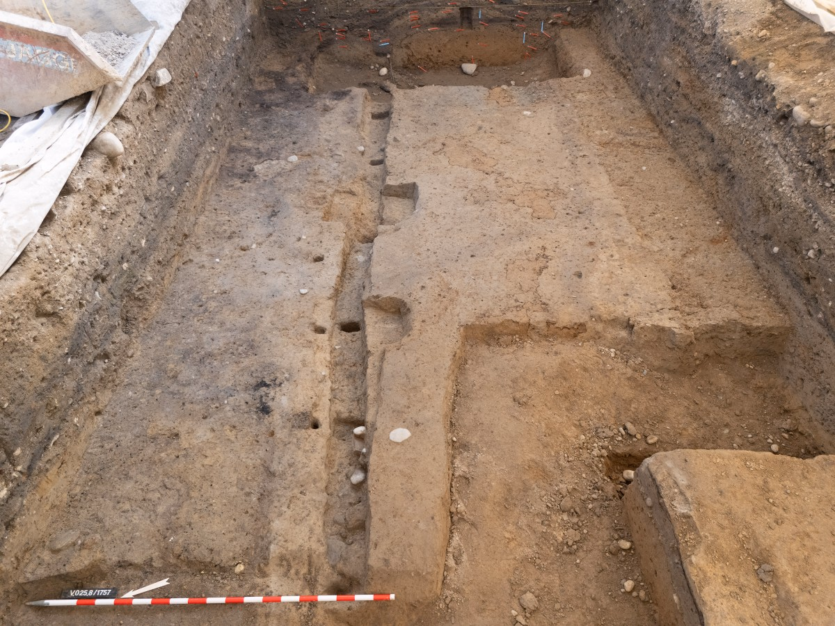 Rechteckiger Ausgrabungsschnitt mit freigelegten, helleren Fundament- oder Bodenspuren; seitliche Profilschnitte zeigen dunklere Schichten, und im Vordergrund liegt ein rot-weiss gerasterter Massstab zur Grössenorientierung.