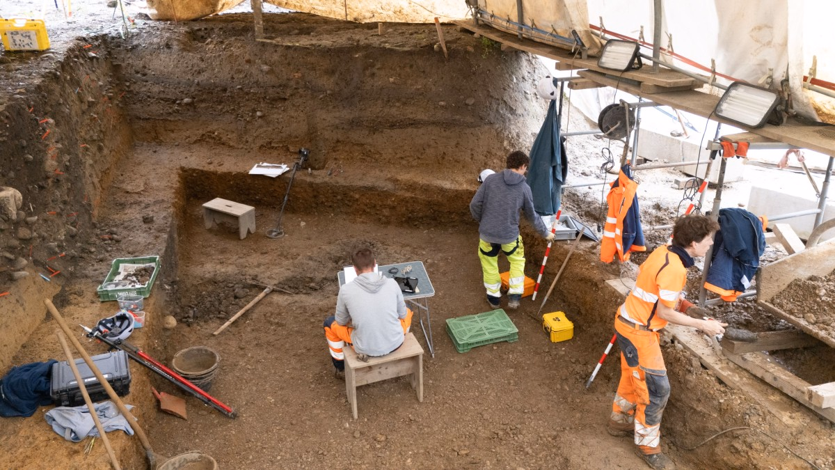 Mehrere Archäologinnen und Archäologen arbeiten in einem mehrstufigen, überdachten Ausgrabungsschnitt, markieren Erdschichten mit roten Stäbchen, sichten Funde in grünen Kisten und notieren ihre Ergebnisse an einem kleinen Klapptisch.