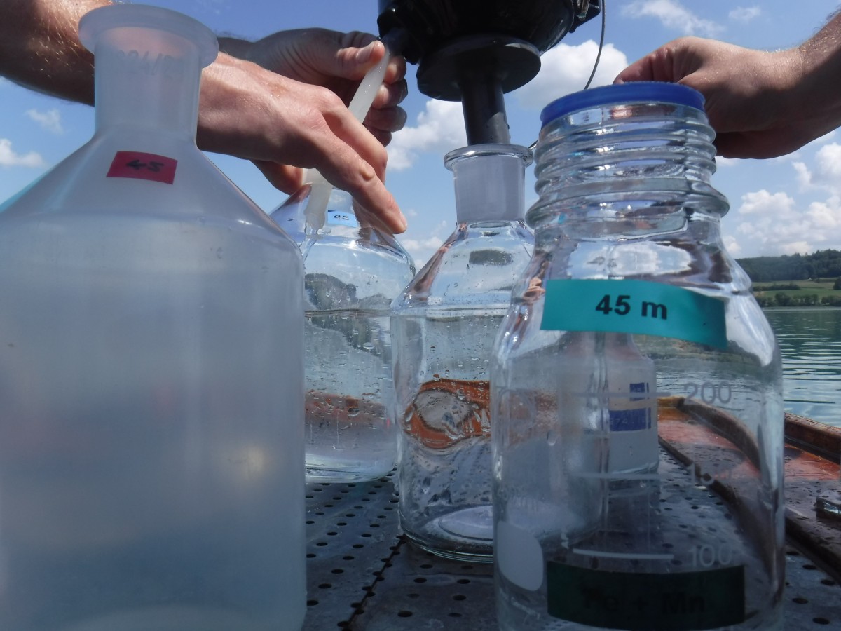 Nahaufnahme von Probenflaschen mit Wasser auf einem Metalltisch, beschriftet mit Farbetiketten, und Händen, die mit einer Pipette arbeiten. Im Hintergrund sind ein See und blauer Himmel sichtbar.