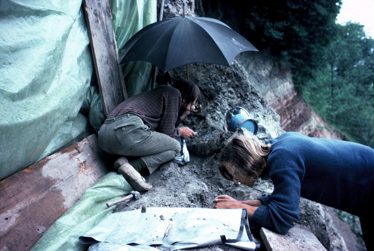 Zwei Personen untersuchen unter einem Regenschirm eine Erdschicht an einer Abbruchkante mit Werkzeugen. Im Vordergrund liegen Planen und Arbeitsmaterialien.