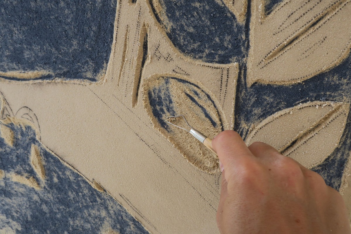 Eine Hand kratzt mit einem Feinwerkzeug eine Textur oder eine Form aus einem beige-blauen festen Material.