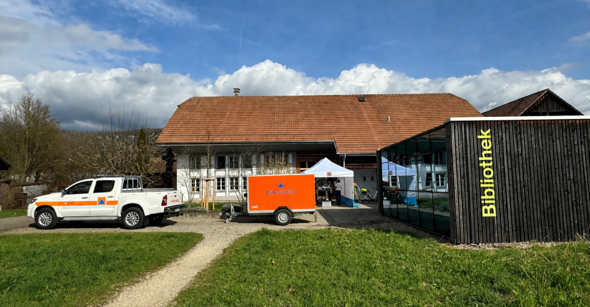 Zwei Fahrzeuge und ein Anhänger der Zivilschutzorganisation vor einem Gebäude mit Bibliothek und Zelt, Wiese und blau bewölktem Himmel.