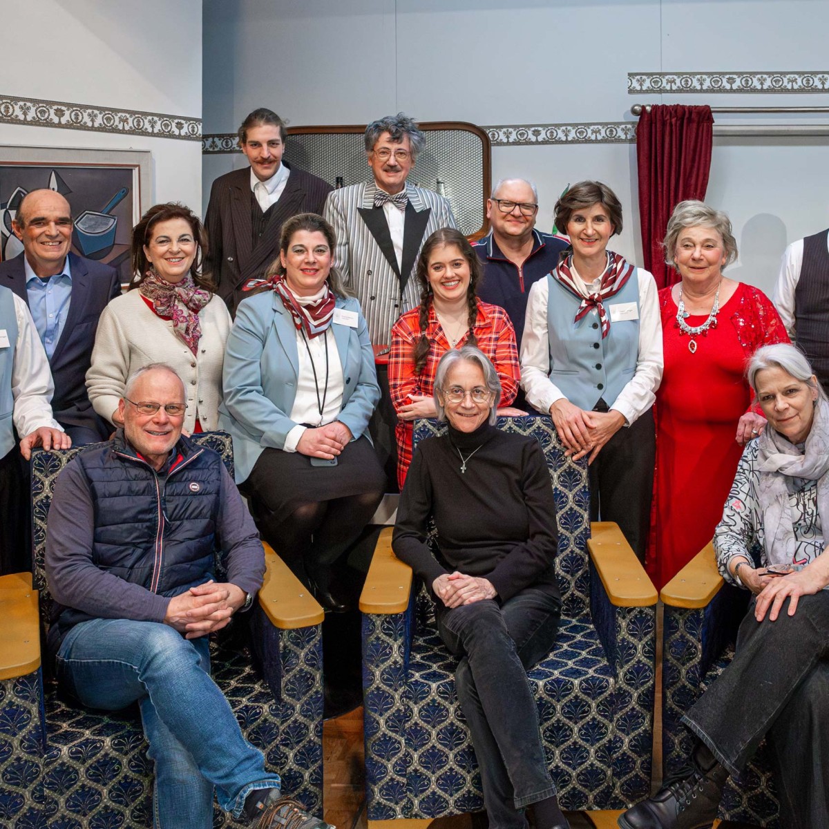 Eine Gruppe Theaterleute stehen zusammen während drei Personen im Vordergrund auf Sitzpolstern mit überschlagenden Beinen Platz genommen haben.