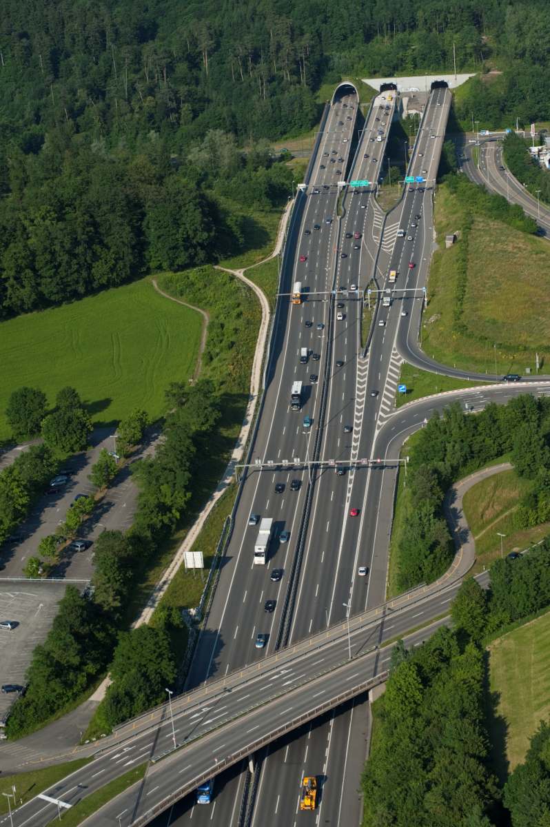Luftaufnahme einer breiten Autobahn mit mehreren Fahrspuren, die durch eine grüne Landschaft zu zwei Tunneln führt. Autos und Lastwagen sind auf der Strecke unterwegs, eine Brücke quert die Fahrbahn im Vordergrund.