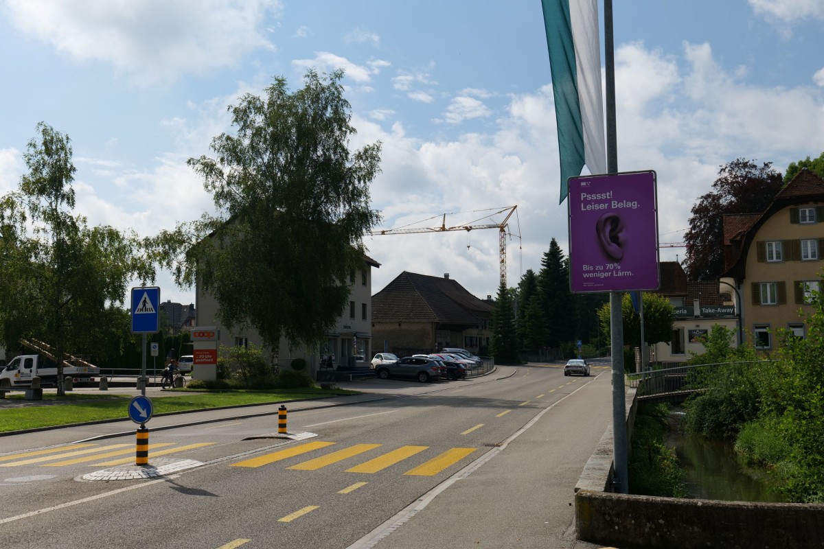 Violett-pinke Infotafel am Strassenrand mit einem abgebildeten Ohr und dem Schriftzug "Psst! Leiser Belag. Bis zu 70% weniger Lärm."