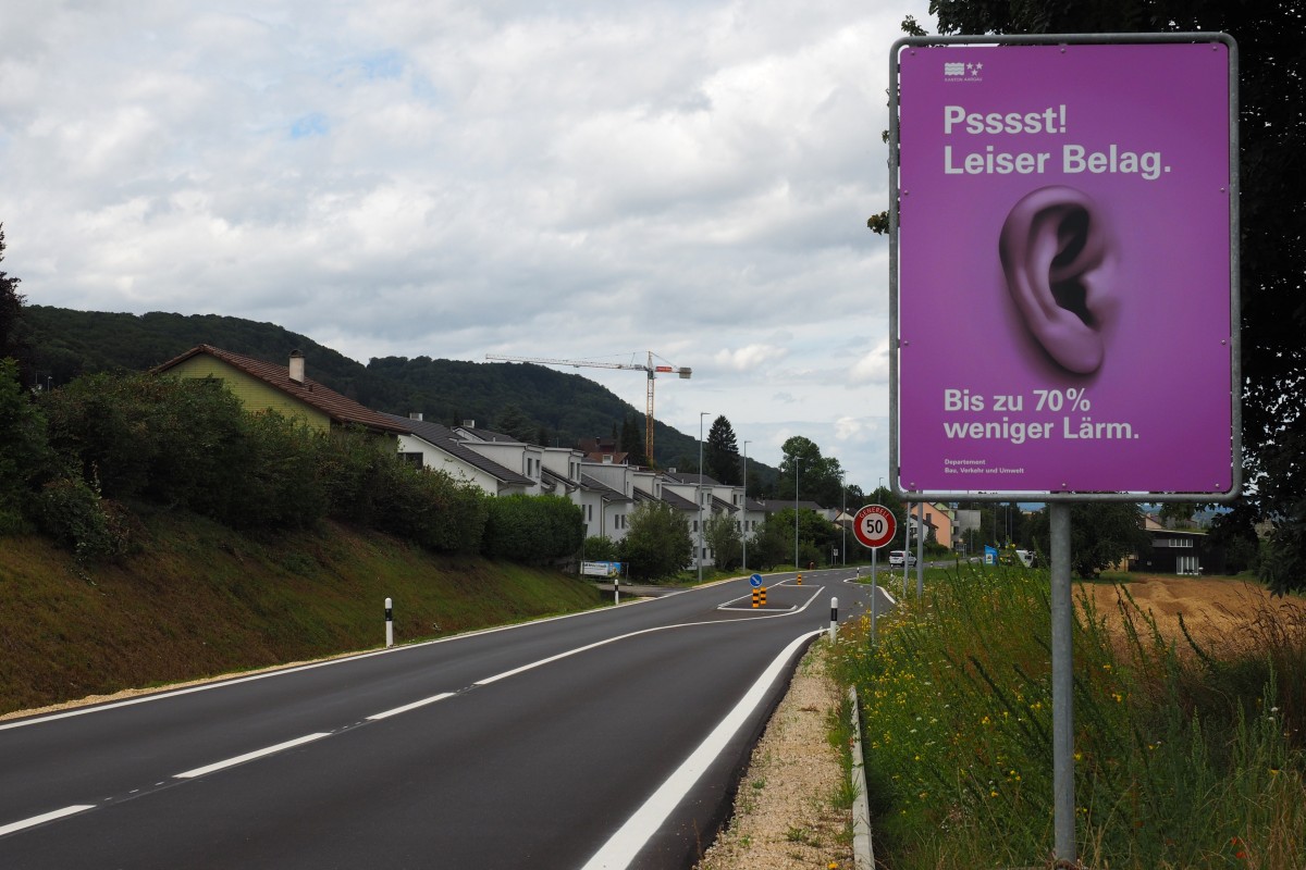 Violett-pinke Infotafel am Strassenrand mit einem abgebildeten Ohr und dem Schriftzug "Psst! Leiser Belag. Bis zu 70% weniger Lärm."