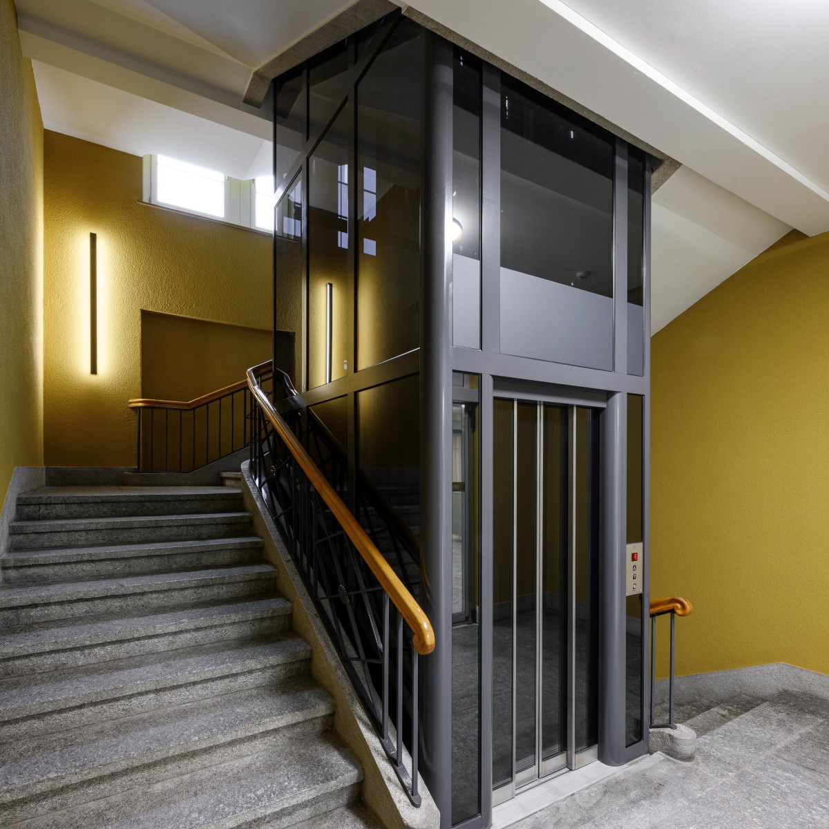 Treppenhaus mit Steintreppen, einem Lift und senfgelben Wänden