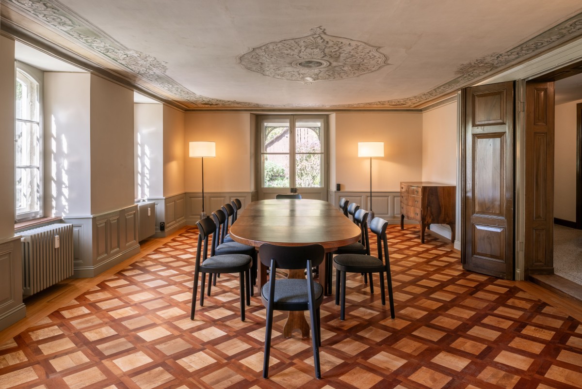 Raum mit Holzboden und filigraner Decke mit Holztisch und Stühlen in der Mitte.