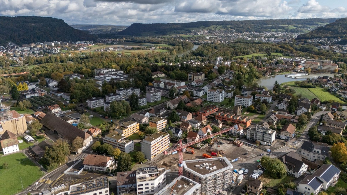 Luftaufnahme eines Stadtteils mit Baustelle und modernen Gebäuden im Vordergrund, umgeben von Wohnhäusern, Grünflächen und einem Fluss. Im Hintergrund Hügel, Wälder und Felder unter bewölktem Himmel.
