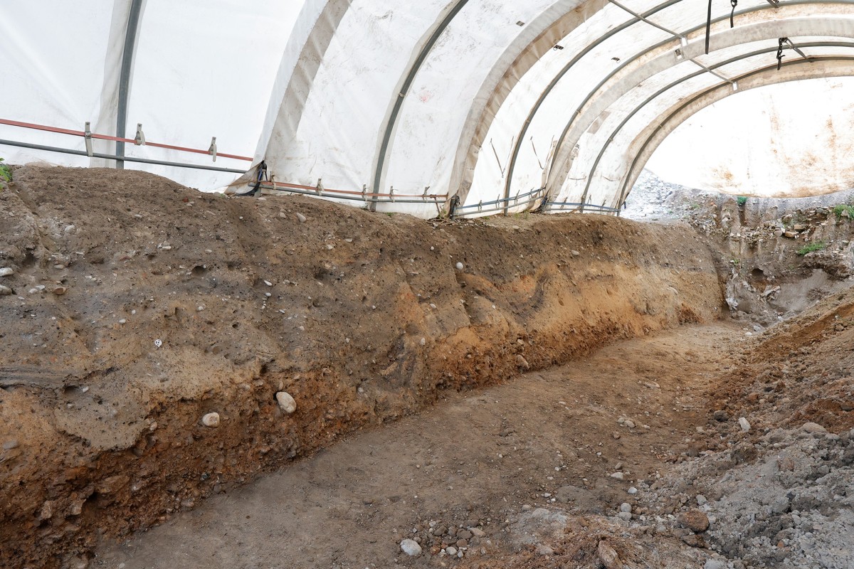 Ein geschützter Grabungsbereich unter einem weissen Zeltdach. Zu sehen ist eine Erdschicht mit sichtbaren Sedimenten und Steinen. Der Boden ist freigelegt, an den Seitenwänden sind unterschiedliche Erdschichten erkennbar.