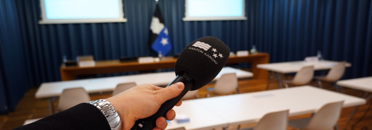 Sicht in den Medienkonferenzraum in Aarau. Zu sehen ist ein Mikrofon, Leinwände und ein Raum mit Stühlen und Tischen.