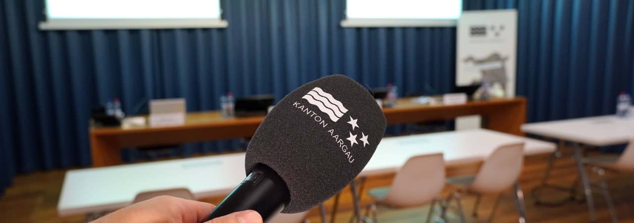 Sicht in den Medienkonferenzraum in Aarau. Zu sehen ist ein Mikrofon, Leinwände und ein Raum mit Stühlen und Tischen.