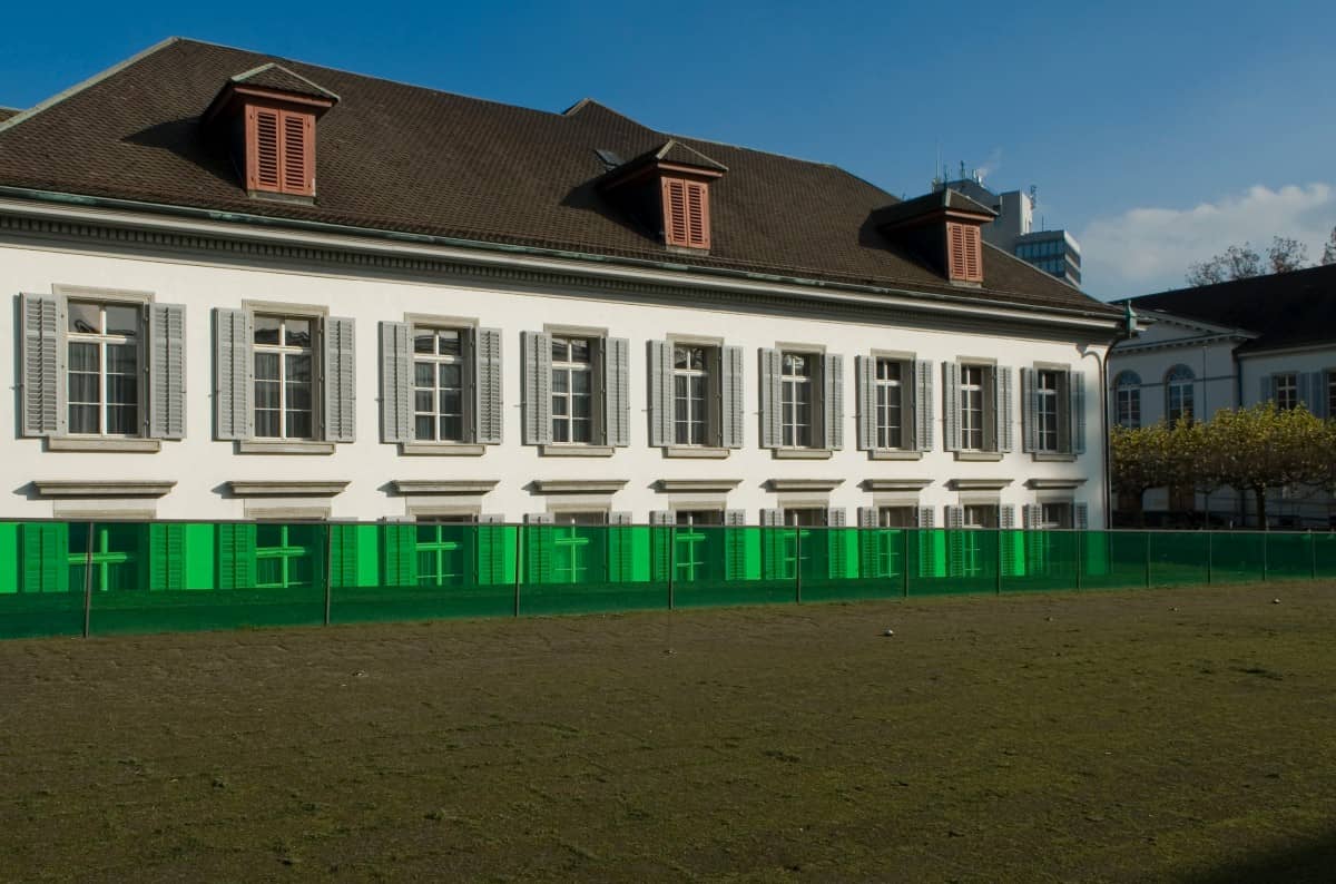 Längsseite des Regierungsgebäudes aus Sicht des Kunsthauses, im Vordergrund brauner Boden, vor dem Gebäude eine kurze, grüne Wand