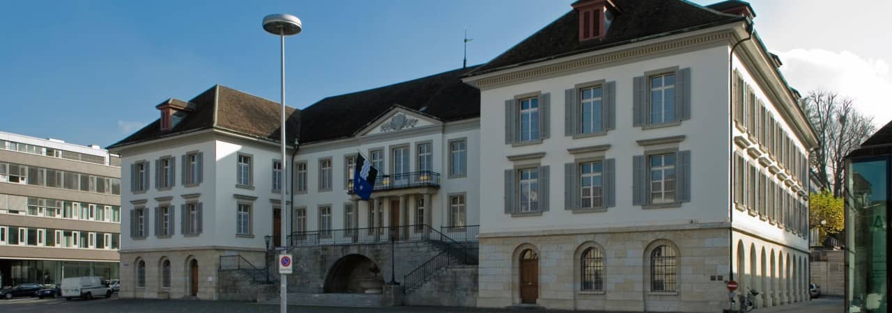 Fotografie des Regierungsgebäudes. Altehrwürdiges Gebäude in Hufeisenform mit einem Balkon in der Mitte mit Aargauer Flagge.