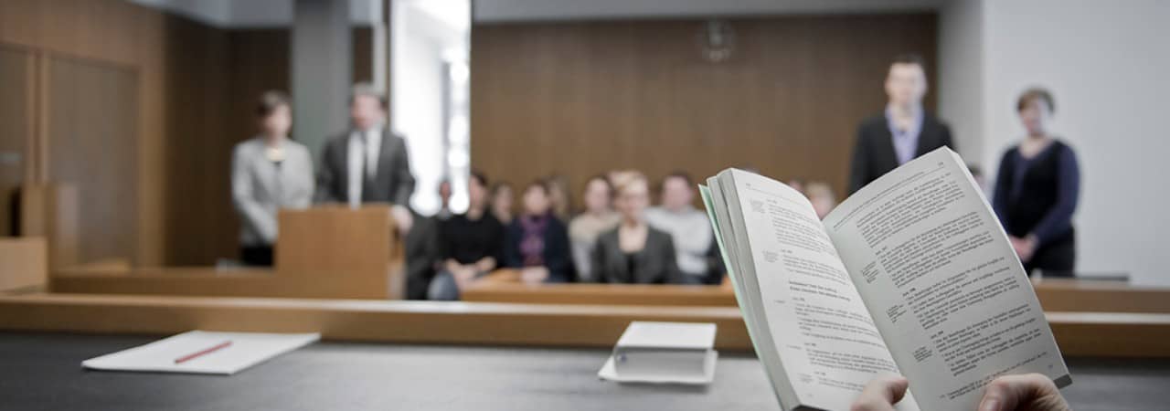 Blick in einen Gerichtssaal mit Personen auf dem Stand und Zuschauern im Hintergrund. Im Vordergrund ein Buch mit Rechtsartikeln.