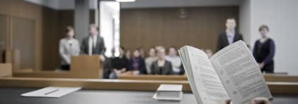 Blick in einen Gerichtssaal mit Personen auf dem Stand und Zuschauern im Hintergrund. Im Vordergrund ein Buch mit Rechtsartikeln.