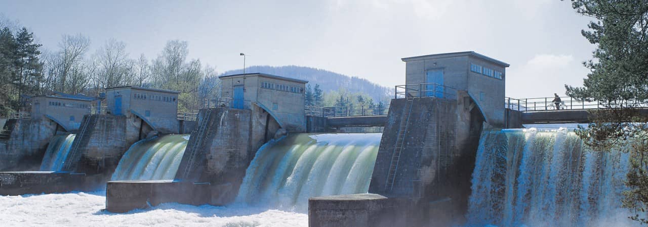 Blick auf ein Wasserkraftwerk. Vier Wasserströme gelangen zwischen vier Mauern mit einem Gebäude darauf durch. Eine Brücke führt über die Wasserfälle. darüber.